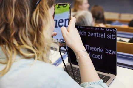 Eine Studentin von hinten zu sehen im Hörsaal, auf dem Tisch ein Laptop mit stark vergrößertem weißem Text auf schwarzem Hintergrund. Sie hält ein Handy in der Hand, auf dem stark vergrößert wenige Buchstaben zu sehen sind.  